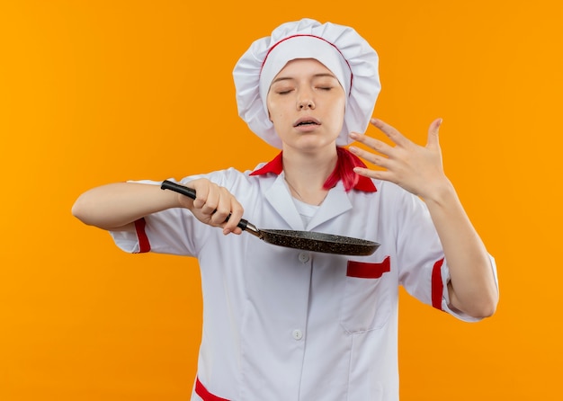 Young pleased blonde female chef in chef uniform holds frying pan and pretends to smell isolated on orange wall