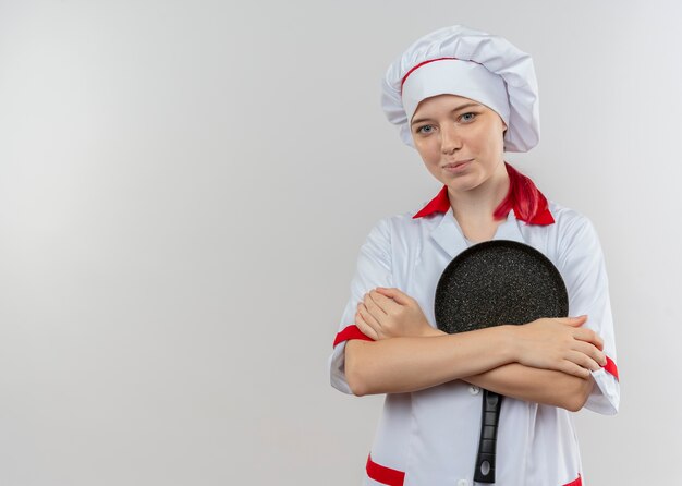 Young pleased blonde female chef in chef uniform holds frying pan with arms isolated on white wall