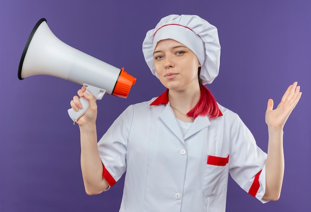 Il giovane chef femmina bionda felice in uniforme da chef tiene l'altoparlante e alza la mano isolata sulla parete viola