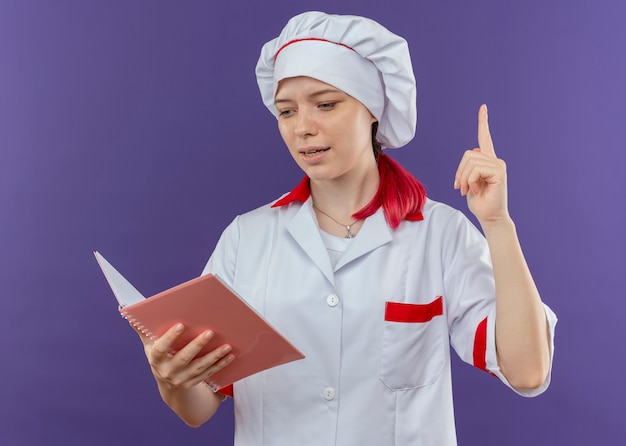 Free photo young pleased blonde female chef in chef uniform holds notebook and points up isolated on violet wall