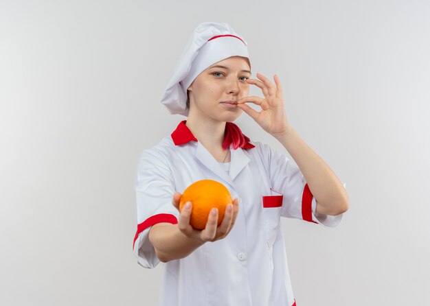 Молодая довольная блондинка-шеф-повар в униформе шеф-повара держит апельсин и жестикулирует вкусный знак, изолированный на белой стене