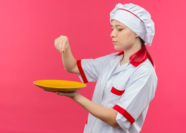 Молодая довольная блондинка-шеф-повар в форме шеф-повара держит тарелку и делает вид, что солит, изолированную на розовой стене