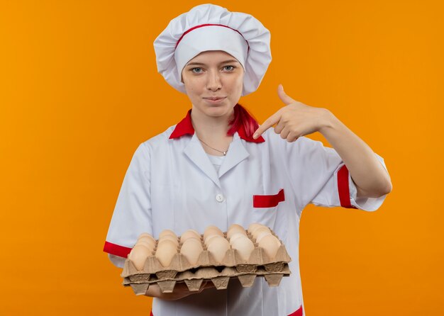 Young pleased blonde female chef in chef uniform holds and points at batch of eggs isolated on orange wall