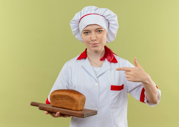 Young pleased blonde female chef in chef uniform holds and points at bread on cutting board isolated on green wall