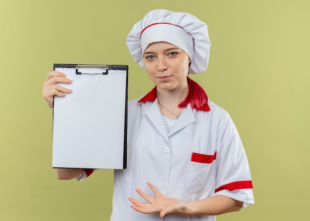 Foto gratuita giovane chef femmina bionda felice in uniforme del cuoco unico tiene e punti negli appunti isolati sulla parete verde