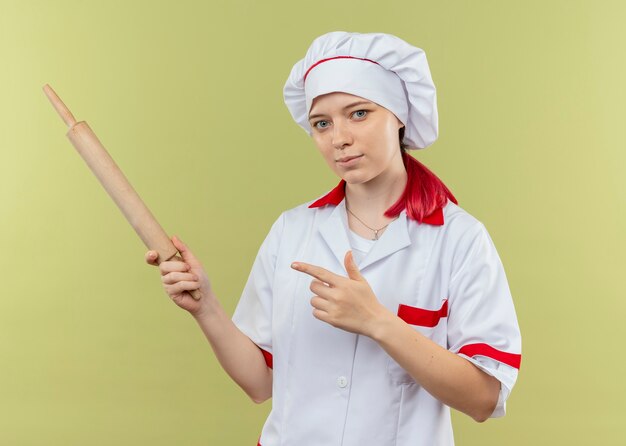 Young pleased blonde female chef in chef uniform holds and points at rolling pin isolated on green wall