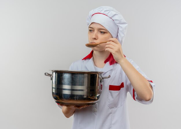 Young pleased blonde female chef in chef uniform holds pot and pretends to try with spoon isolated on white wall