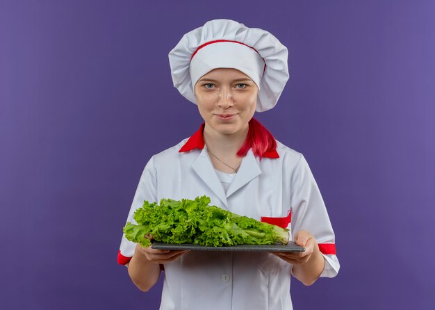 Молодая довольная блондинка-шеф-повар в форме шеф-повара держит салат на разделочной доске, изолированной на фиолетовой стене