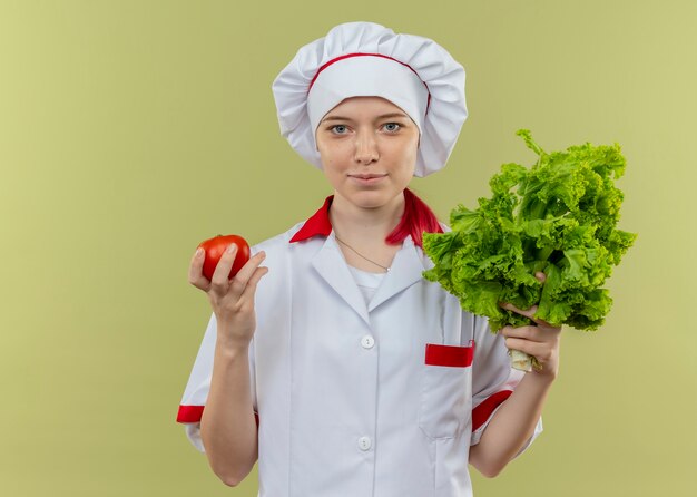 Young pleased blonde female chef in chef uniform holds tomato and salad isolated on green wall