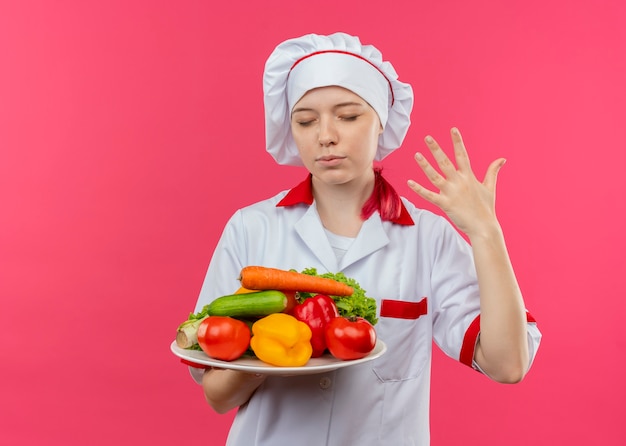 Il giovane chef femmina bionda felice in uniforme da chef tiene le verdure sul piatto e solleva la mano isolata sulla parete rosa