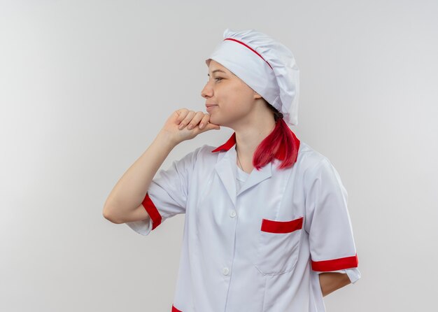 Молодая довольная блондинка-шеф-повар в униформе смотрит в сторону и кладет руку на подбородок, изолированную на белой стене