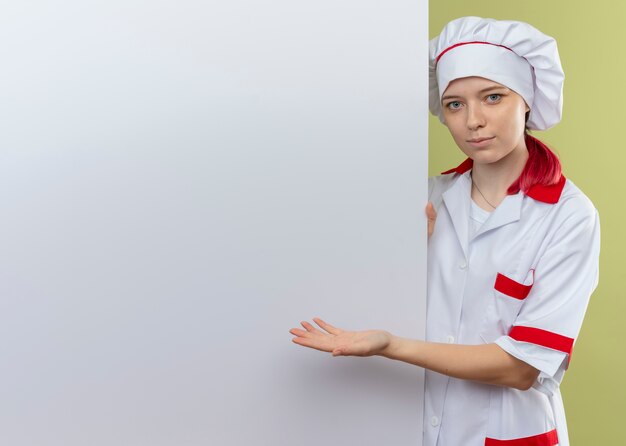 Young pleased blonde female chef in chef uniform stands behind and points at white wall isolated on green wall