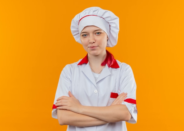 Il giovane chef femmina bionda felice in uniforme da chef sta con le braccia incrociate isolato sulla parete arancione