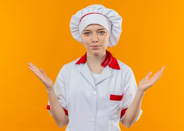 Young pleased blonde female chef in chef uniform stands with open hands isolated on orange wall