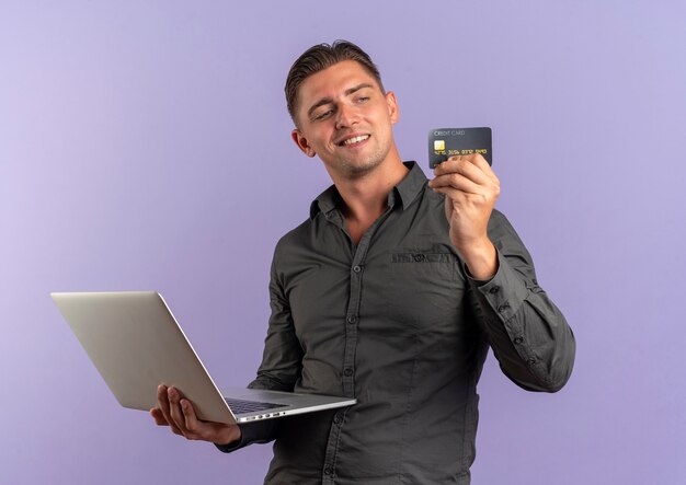 young pleased blonde handsome man holds laptop and looks at credit card