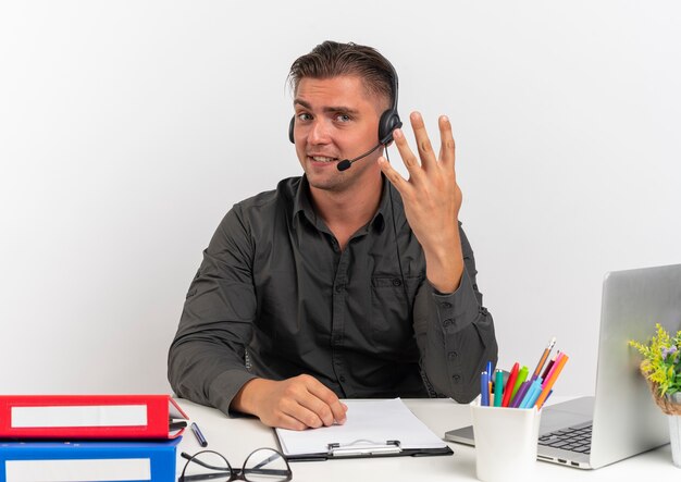 Молодой довольный блондин офисный работник человек в наушниках сидит за столом с офисными инструментами, используя жесты ноутбука четыре пальцами