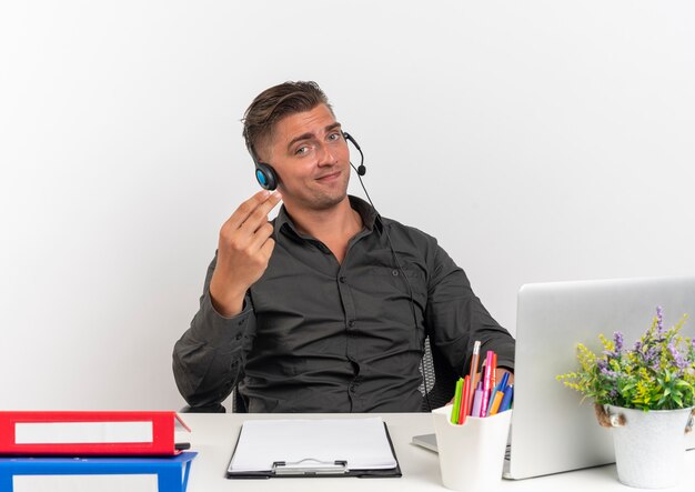 Young pleased blonde office worker man on headphones sits at desk with office tools using laptop gestures money hand sign isolated on white background with copy space