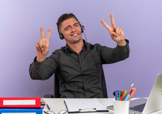 Young pleased blonde office worker man on headphones sits at desk with office tools using laptop gestures victory hand sign isolated on violet background with copy space