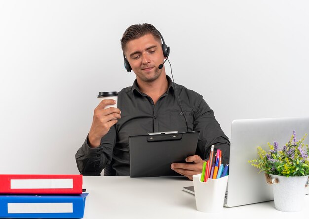 Young pleased blonde office worker man on headphones sits at desk with office tools using laptop holds clipboard and coffee cup isolated on white background with copy space