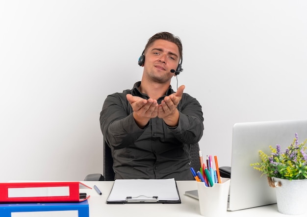 L'uomo di lavoratore di ufficio bionda felice giovane sulle cuffie si siede alla scrivania con strumenti di ufficio utilizzando il computer portatile tiene le mani insieme isolato su sfondo bianco con spazio di copia