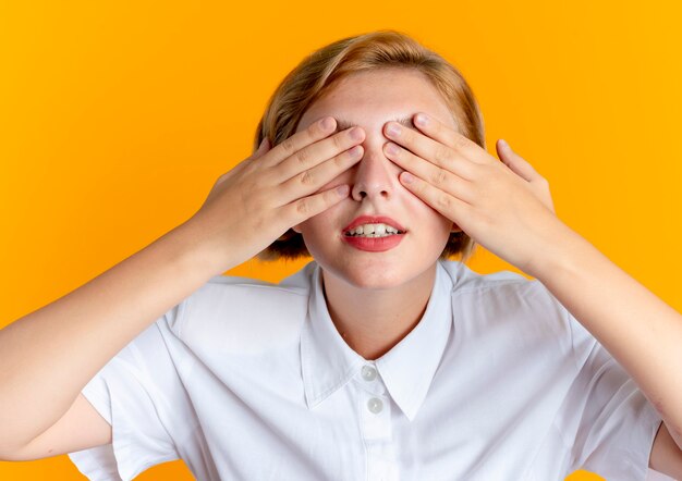 Young pleased blonde russian girl closes eyes with hands isolated on orange background with copy space