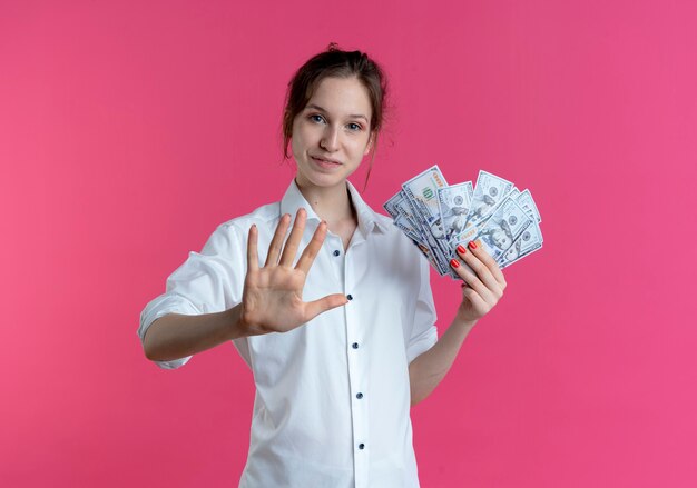 Молодая довольная русская блондинка жестами пятерых держит деньги на розовом с копией пространства