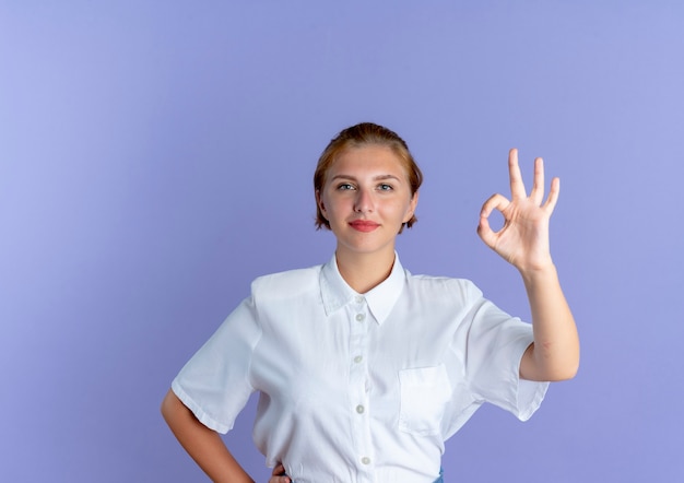 Young pleased blonde russian girl gestures ok hand sign isolated on purple background with copy space
