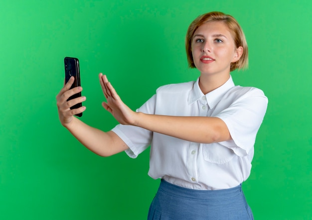 Young pleased blonde russian girl holds phone with raised hand and looks at side isolated on green background with copy space