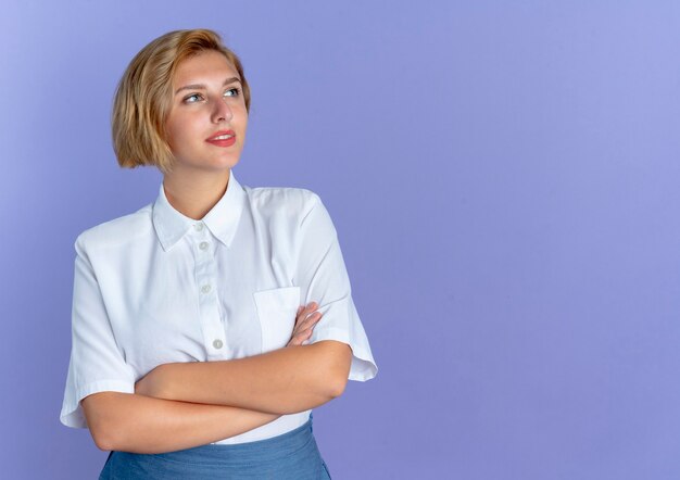 Young pleased blonde russian girl looks at side with crossed arms isolated on purple background with copy space