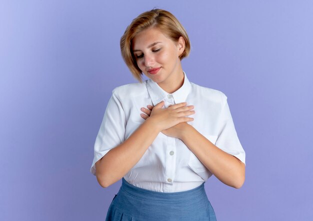 Free photo young pleased blonde russian girl puts hands on chest looking down isolated on purple background with copy space