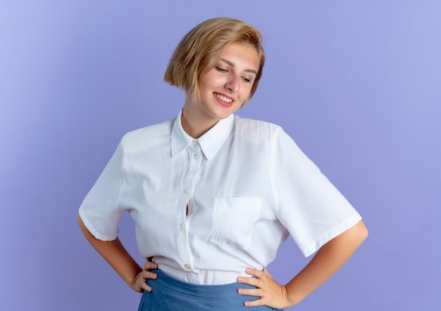 Foto gratuita la giovane ragazza russa bionda lieta mette le mani sulla vita che osserva giù isolata su fondo viola con lo spazio della copia