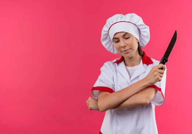 Young pleased caucasian cook girl in chef uniform crosses arms and holds knife isolated on pink background with copy space