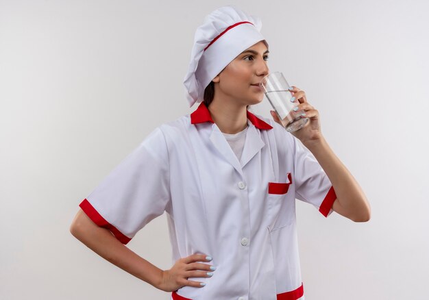 Young pleased caucasian cook girl in chef uniform drinks glass of water on white  with copy space