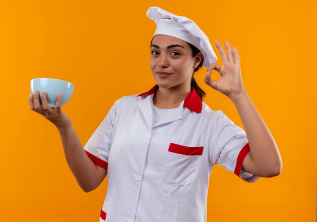 Young pleased caucasian cook girl in chef uniform holds bowl and gestures ok hand sign isolated on orange wall with copy space