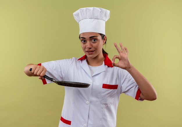 Молодая довольная кавказская девушка-повар в униформе шеф-повара держит сковороду и жестами показывает знак рукой на зеленой стене с копией пространства