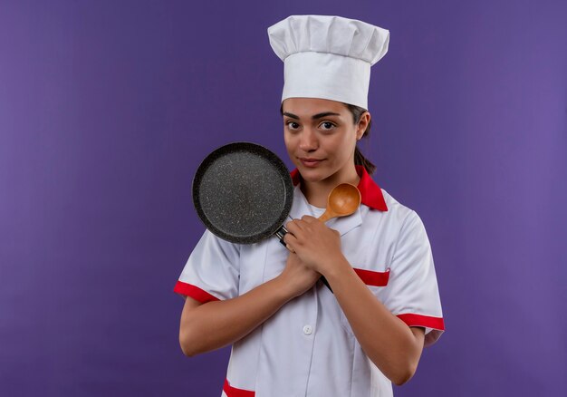 La giovane ragazza caucasica felice del cuoco in uniforme del cuoco unico tiene la padella e il cucchiaio di legno isolati sulla parete viola con lo spazio della copia