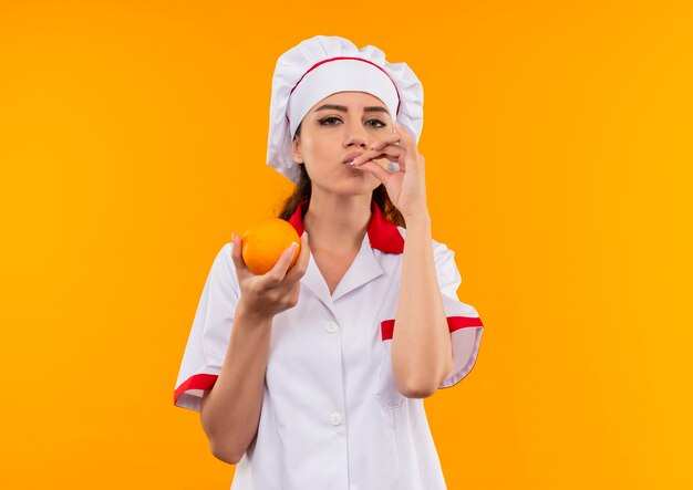 Young pleased caucasian cook girl in chef uniform holds orange and gestures tasty delicious sign isolated on orange wall with copy space