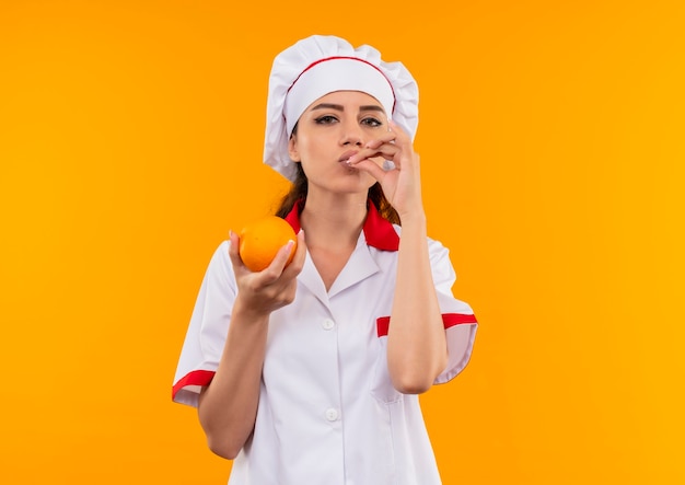 Young pleased caucasian cook girl in chef uniform holds orange and gestures tasty delicious sign isolated on orange wall with copy space