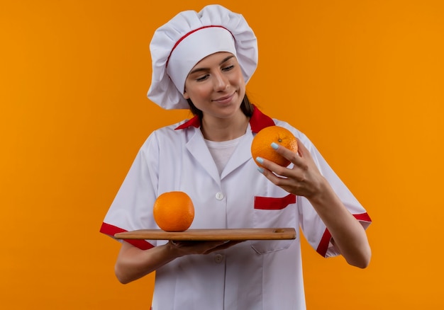 요리사 제복을 입은 젊은 기쁘게 백인 요리사 소녀 커팅 보드에 오렌지를 보유하고 손에 복사 공간이있는 오렌지 공간에 고립 된