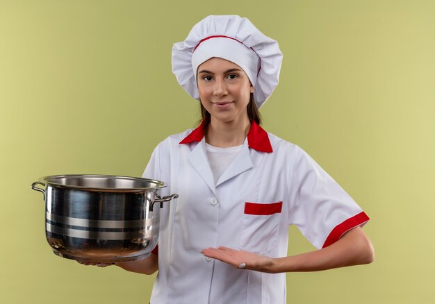 Young pleased caucasian cook girl in chef uniform holds and points at pot isolated on green background with copy space