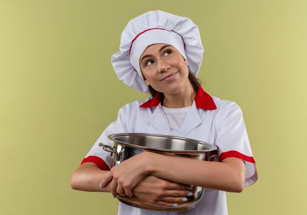 Young pleased caucasian cook girl in chef uniform holds pot with arms and looks at side on green  with copy space