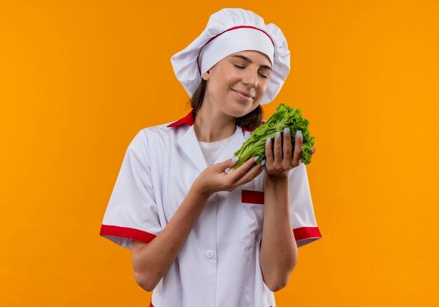 Young pleased caucasian cook girl in chef uniform holds and pretends to smell salad isolated on orange space with copy space