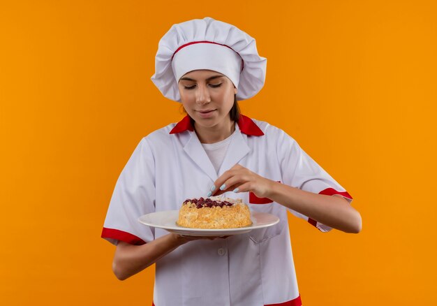 Young pleased caucasian cook girl in chef uniform holds and pretends to try cake on plate  isolated on orange space with copy space