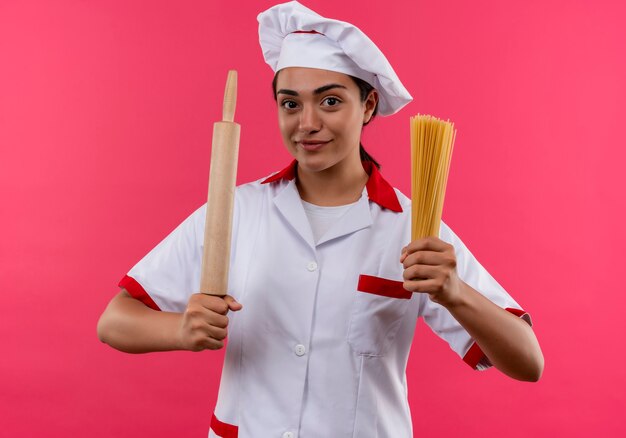 シェフの制服を着た若い喜んでいる白人料理人の女の子は、コピースペースでピンクの壁に分離された麺棒とスパゲッティの束を保持します