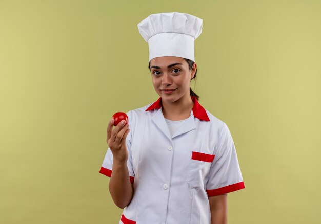 Young pleased caucasian cook girl in chef uniform holds tomato isolated on green wall with copy space