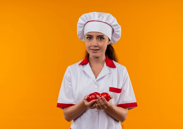 Young pleased caucasian cook girl in chef uniform holds tomatoes in hands isolated on orange wall with copy space