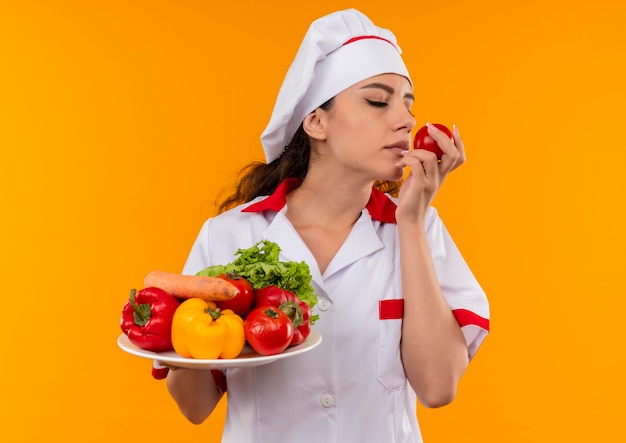 La giovane ragazza caucasica felice del cuoco in uniforme del cuoco unico tiene le verdure sul piatto e finge di annusare il pomodoro isolato sulla parete arancione con lo spazio della copia