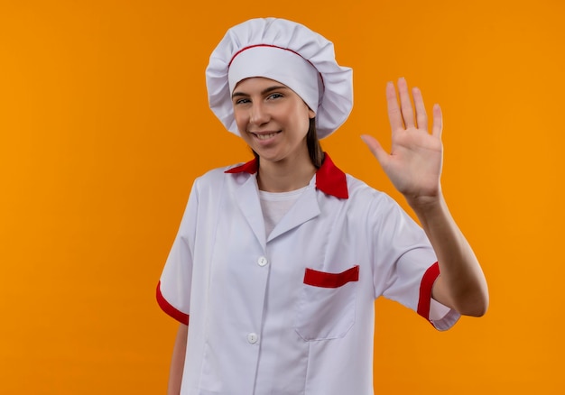 Young pleased caucasian cook girl in chef uniform raises hand on orange  with copy space