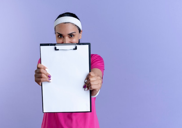 Young pleased caucasian sporty woman wearing headband and wristbands holds out clipboard isolated on purple space with copy space