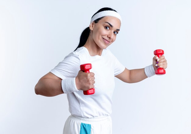 Young pleased caucasian sporty woman wearing headband and wristbands stands sideways holding dumbbells isolated on white space with copy space